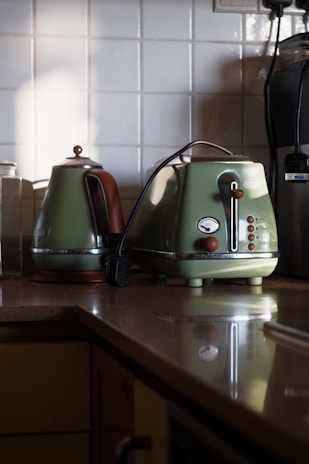 A kitchen countertop featuring a classic green kettle and toaster set. The countertop is made of a smooth, reflective surface. The kettle has a brown handle and a polished metal spout, while the toaster has round buttons and a dial. The background consists of white tiled walls and a few additional kitchen appliances partially visible.