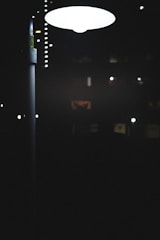 A streetlamp emits a bright, white light in a dark, nighttime setting. Various smaller lights and faintly illuminated windows appear in the distance, suggesting an urban environment.