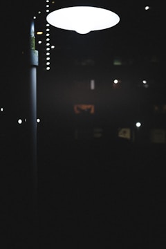 A streetlamp emits a bright, white light in a dark, nighttime setting. Various smaller lights and faintly illuminated windows appear in the distance, suggesting an urban environment.