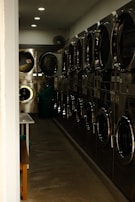 A narrow hallway of a laundromat with several stainless steel washing machines lined up on one side. The lighting is dim, casting shadows along the floor and machines. A few ceiling lights illuminate the space, and a folding table and bench are partially visible on the left. In the back, a basket is visible on the floor.