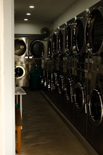 A narrow hallway of a laundromat with several stainless steel washing machines lined up on one side. The lighting is dim, casting shadows along the floor and machines. A few ceiling lights illuminate the space, and a folding table and bench are partially visible on the left. In the back, a basket is visible on the floor.