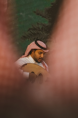 A man is playing an oud while wearing traditional Middle Eastern attire, including a white thobe and a red-checkered keffiyeh. The image is framed by a blurred foreground, giving a sense of depth and focus on the musician. The background appears to be a natural setting with dark green foliage.