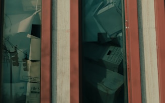 A close-up view of a window showing a collection of file boxes and documents stacked behind the glass. The window frame is vertical with a base color that appears to be orange or red paired with beige trim. The file boxes are labeled and variously stacked, giving the appearance of storage or archiving.