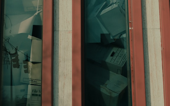 A close-up view of a window showing a collection of file boxes and documents stacked behind the glass. The window frame is vertical with a base color that appears to be orange or red paired with beige trim. The file boxes are labeled and variously stacked, giving the appearance of storage or archiving.