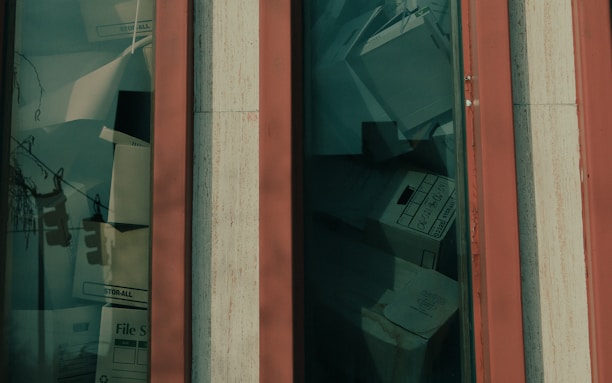 A close-up view of a window showing a collection of file boxes and documents stacked behind the glass. The window frame is vertical with a base color that appears to be orange or red paired with beige trim. The file boxes are labeled and variously stacked, giving the appearance of storage or archiving.