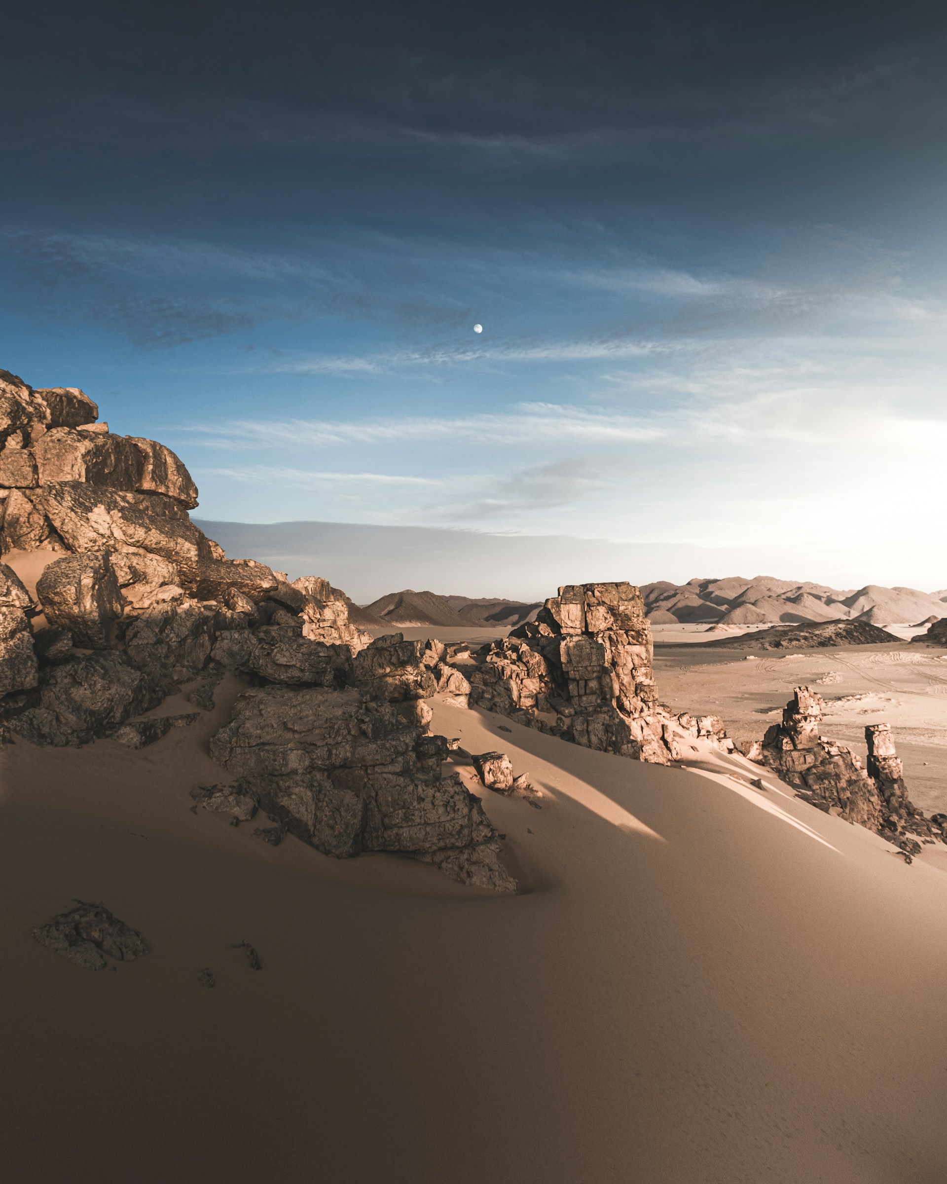 A wide-open desert landscape under a vast blue sky, with rugged rock formations casting long shadows.