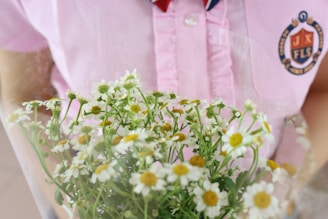 A bouquet of small white flowers with yellow centers is held against a pink shirt with a crest on the left side.
