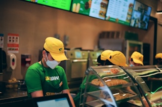 People wearing green uniforms and yellow caps are working behind a counter at a fast-food restaurant. A menu is displayed on digital screens above, and there is a clear display case with baked goods in the foreground.