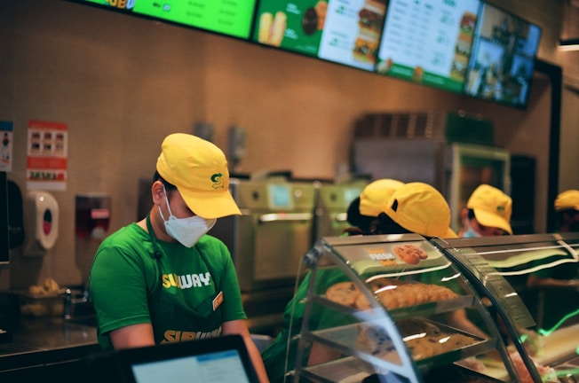 People wearing green uniforms and yellow caps are working behind a counter at a fast-food restaurant. A menu is displayed on digital screens above, and there is a clear display case with baked goods in the foreground.