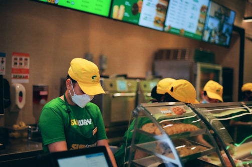 People wearing green uniforms and yellow caps are working behind a counter at a fast-food restaurant. A menu is displayed on digital screens above, and there is a clear display case with baked goods in the foreground.