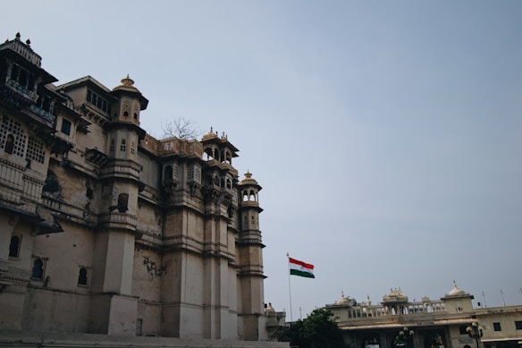 A grand, historical building with intricate architectural details and multiple levels of windows and balconies. The structure seems aged, with some visible wear on the walls. In the background, an Indian flag flies high atop a flagpole, adding color to the muted tones of the structure. The sky is overcast, adding a somber tone to the scene.