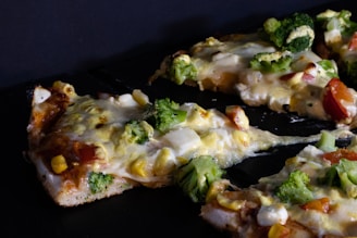 Slices of pizza are topped with vegetables such as broccoli, corn, and tomatoes, with melted cheese visibly covering the toppings. The crust appears to be golden brown and slightly thick, with a homemade appearance.
