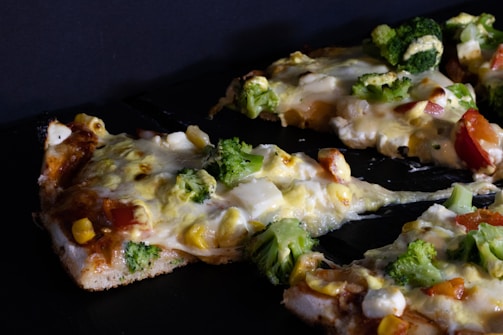 Slices of pizza are topped with vegetables such as broccoli, corn, and tomatoes, with melted cheese visibly covering the toppings. The crust appears to be golden brown and slightly thick, with a homemade appearance.