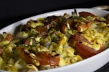 A close-up of a baked dish featuring melted cheese, sliced tomatoes, and herbs sprinkled on top. The colors are rich and the dish appears to be hot and freshly prepared in a white ceramic baking dish.
