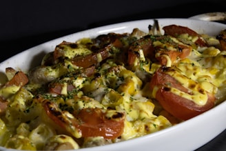 A close-up of a baked dish featuring melted cheese, sliced tomatoes, and herbs sprinkled on top. The colors are rich and the dish appears to be hot and freshly prepared in a white ceramic baking dish.