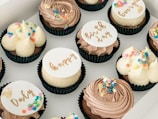 A selection of artisanal cupcakes arranged in a grid pattern within a white box. Each cupcake features different frosting styles and is adorned with colorful sprinkles or ornate inscriptions on fondant discs.