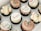 A selection of artisanal cupcakes arranged in a grid pattern within a white box. Each cupcake features different frosting styles and is adorned with colorful sprinkles or ornate inscriptions on fondant discs.