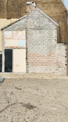 An exposed section of a brick wall reveals different stages of construction and materials, including concrete blocks and various types of bricks. The wall varies in color and texture, showing areas of wear and some signs of decay. The ground in front appears bare and unfinished, with a rough surface.