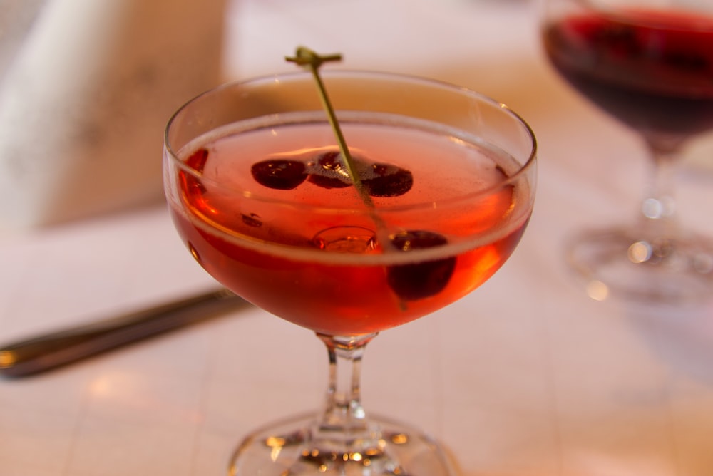 a glass of red liquid with a green stem and a black olive on top