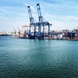 Large industrial cranes dominate a bustling port where numerous shipping containers are stacked, ready for transport. The scene is set against a backdrop of ships, cranes, and a distant city skyline under a partly cloudy blue sky. The calm water in the foreground reflects the industrial activity.