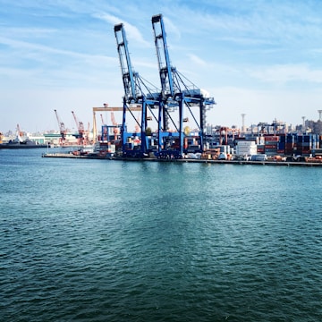 Large industrial cranes dominate a bustling port where numerous shipping containers are stacked, ready for transport. The scene is set against a backdrop of ships, cranes, and a distant city skyline under a partly cloudy blue sky. The calm water in the foreground reflects the industrial activity.