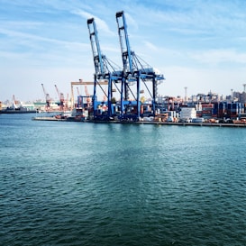 Large industrial cranes dominate a bustling port where numerous shipping containers are stacked, ready for transport. The scene is set against a backdrop of ships, cranes, and a distant city skyline under a partly cloudy blue sky. The calm water in the foreground reflects the industrial activity.