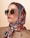 A woman wearing oversized square sunglasses and a colorful silk scarf around her head. The scarf features a vibrant pattern with shades of red, blue, and beige. She is dressed in a beige outfit with a subtle zipper detail.