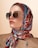 A woman wearing oversized square sunglasses and a colorful silk scarf around her head. The scarf features a vibrant pattern with shades of red, blue, and beige. She is dressed in a beige outfit with a subtle zipper detail.