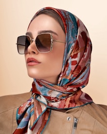 A woman wearing oversized square sunglasses and a colorful silk scarf around her head. The scarf features a vibrant pattern with shades of red, blue, and beige. She is dressed in a beige outfit with a subtle zipper detail.