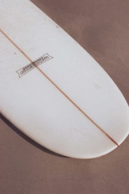 A close-up view of the tail end of a surfboard resting on a sandy surface. The surfboard has a white color with a single wooden stringer running down the middle. There is a logo printed on the upper left portion.