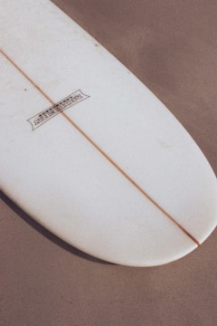 A close-up view of the tail end of a surfboard resting on a sandy surface. The surfboard has a white color with a single wooden stringer running down the middle. There is a logo printed on the upper left portion.
