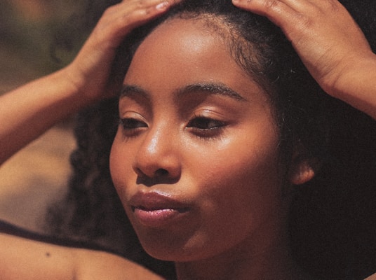 A close-up of a young woman with her hands on her head, showcasing her curly hair and smooth skin. The lighting creates a warm and natural look, highlighting her serene expression.