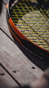 A close-up view of a tennis racket with a bright orange frame and yellow strings. The racket is resting on a wooden surface, showing the texture of the wood with visible screws.