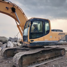 A large yellow excavator with a hydraulic arm is parked on a rocky and muddy terrain. A person is inside the cabin, which is enclosed with glass windows. The background is cloudy, suggesting an overcast day.