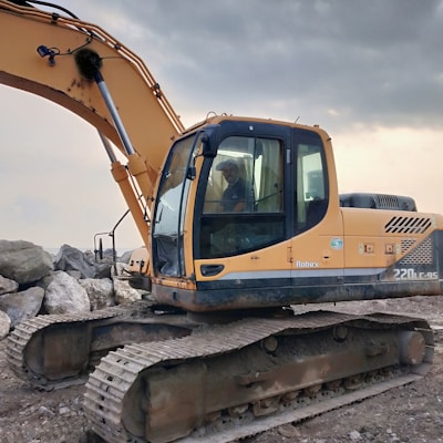 A large yellow excavator with a hydraulic arm is parked on a rocky and muddy terrain. A person is inside the cabin, which is enclosed with glass windows. The background is cloudy, suggesting an overcast day.