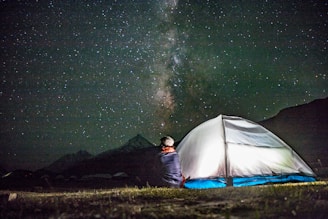 A person is sitting outside near an illuminated tent, gazing at a starry night sky with a visible Milky Way. The scene is set in a natural outdoor environment, likely a remote or mountainous area with dark skies ideal for stargazing.