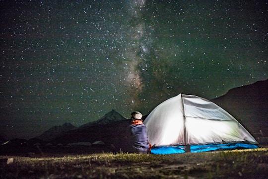 A person is sitting outside near an illuminated tent, gazing at a starry night sky with a visible Milky Way. The scene is set in a natural outdoor environment, likely a remote or mountainous area with dark skies ideal for stargazing.