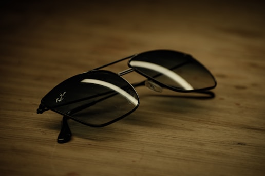 A pair of aviator-style sunglasses with a reflective surface lies on a wooden table. The ambient lighting casts a soft shadow and highlights the sleek, metallic frame of the glasses.