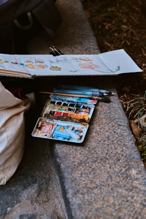 A sketchbook and a watercolor set are placed on the ground next to a stone edge. The sketchbook contains unfinished sketches with some color, depicting various shapes. The watercolor palette is well-used, with multiple colors visible, and a couple of watercolor brushes are resting on top or nearby.