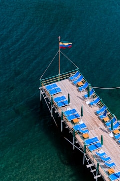 A wooden dock extends over a body of deep, blue-green water. The dock is lined with blue lounge chairs, some of which have orange towels placed on them, and green umbrellas. A flagpole stands at one end of the dock, displaying a tricolor flag with horizontal stripes of yellow, blue, and red.