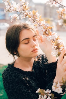 A young woman with closed eyes is standing near a branch of blossoming flowers, gently touching them with her hand. The sunlight casts a warm glow on her face, creating a serene and peaceful atmosphere.