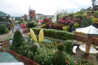 A vibrant and meticulously maintained garden filled with lush greenery, colorful flowers, and decorative sculptures. There is a whimsical building resembling a small tower with a windmill on top, surrounded by diverse plants. Paths wind throughout the garden, with benches for relaxing and an umbrella-shaped structure providing shade. People are casually exploring the garden.
