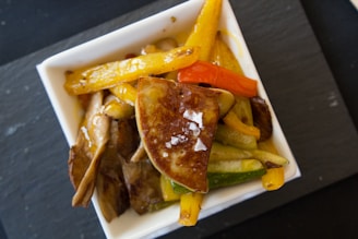 A close-up view of a square white dish filled with a variety of cooked vegetables and sliced mushrooms. The centerpiece appears to be a piece of seared foie gras topped with coarse salt. Yellow and red bell peppers, as well as zucchini slices, are visible, all cooked to a slightly caramelized finish.