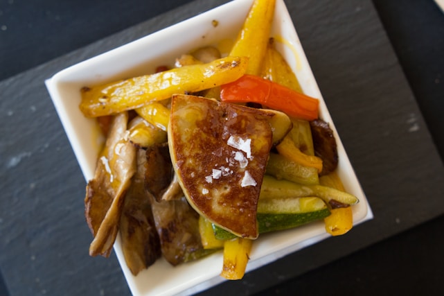 A close-up view of a square white dish filled with a variety of cooked vegetables and sliced mushrooms. The centerpiece appears to be a piece of seared foie gras topped with coarse salt. Yellow and red bell peppers, as well as zucchini slices, are visible, all cooked to a slightly caramelized finish.