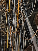 A complex network of illuminated cables and wires suspended from the ceiling, displaying a mix of orange and white glows. The cables intertwine and loop in various directions against a backdrop of a modern industrial interior featuring exposed beams and metal structures.