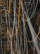 A complex network of illuminated cables and wires suspended from the ceiling, displaying a mix of orange and white glows. The cables intertwine and loop in various directions against a backdrop of a modern industrial interior featuring exposed beams and metal structures.
