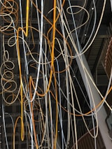 A complex network of illuminated cables and wires suspended from the ceiling, displaying a mix of orange and white glows. The cables intertwine and loop in various directions against a backdrop of a modern industrial interior featuring exposed beams and metal structures.