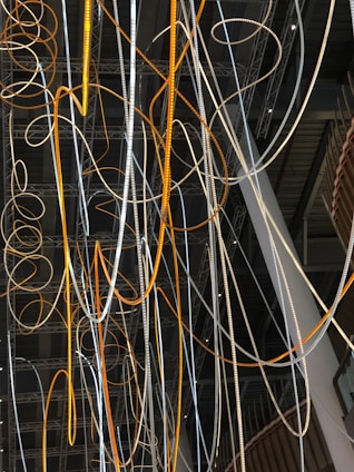 A complex network of illuminated cables and wires suspended from the ceiling, displaying a mix of orange and white glows. The cables intertwine and loop in various directions against a backdrop of a modern industrial interior featuring exposed beams and metal structures.