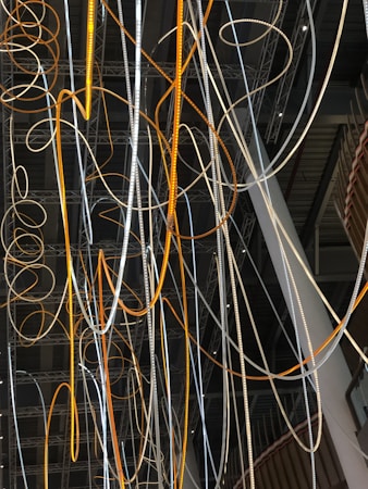 A complex network of illuminated cables and wires suspended from the ceiling, displaying a mix of orange and white glows. The cables intertwine and loop in various directions against a backdrop of a modern industrial interior featuring exposed beams and metal structures.