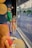 Three mannequins are dressed in summer clothing, including sunglasses, shorts, and hats, displayed in a storefront window. The window reflects a view of a pedestrian street lined with buildings and trees.